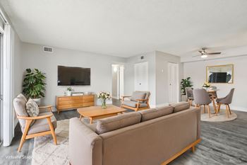 A living room with a brown sofa, wooden chairs, and a tv on the wall